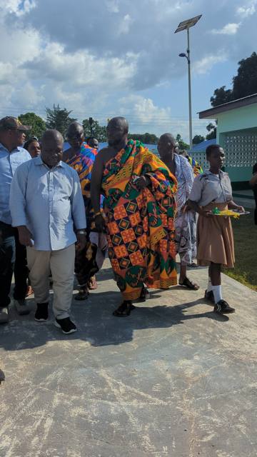 Guests and chiefs walk across the school compound, led by a pupil carrying a tray.