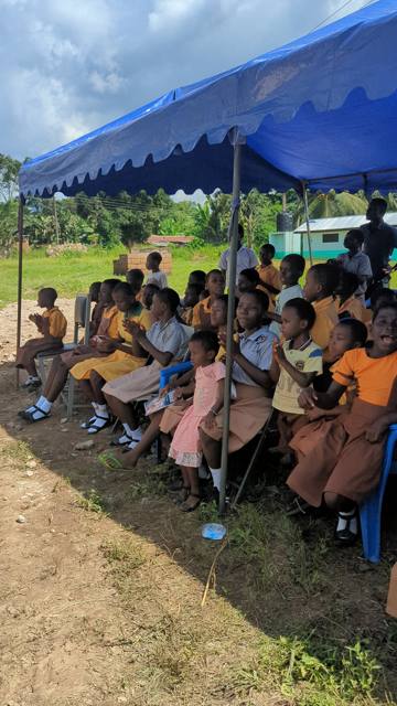 Pupils seated in the shade, applauding.