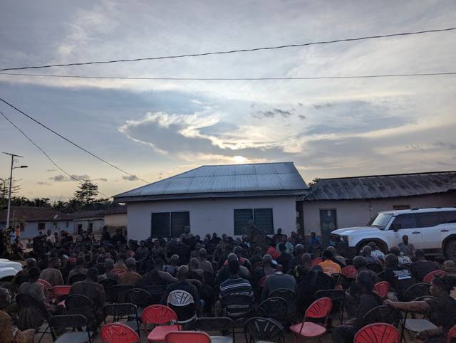The gathering at dusk, seated in rows before the compound.