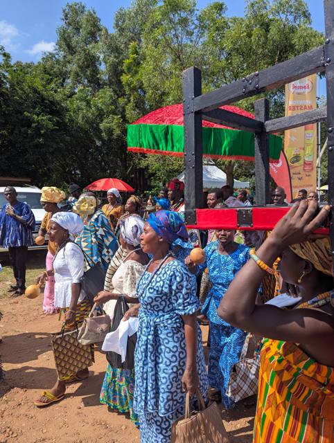 Women carrying gourd rattles move through the festival grounds beside a carried stool.