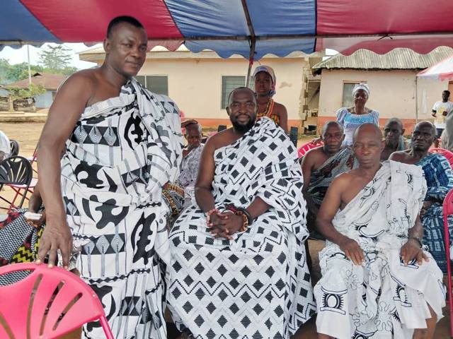 A standing elder beside seated chiefs in black-and-white patterned cloth.